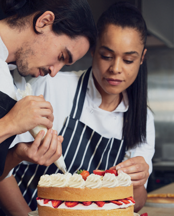 two chefs icing a sponge cake with cream and sliced strawberries